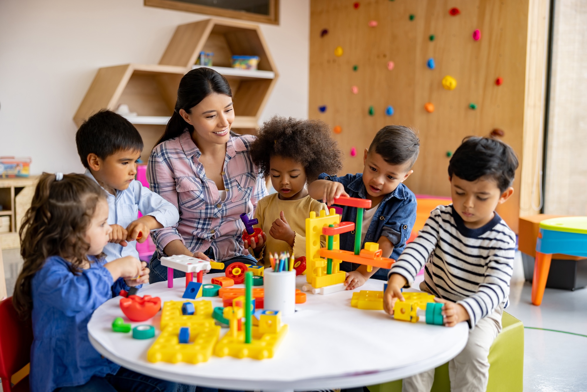 A classroom setting with several students and a teacher sitting around a table. The students are engaged in an activity involving rulers, colored pencils, and paper with geometric designs.